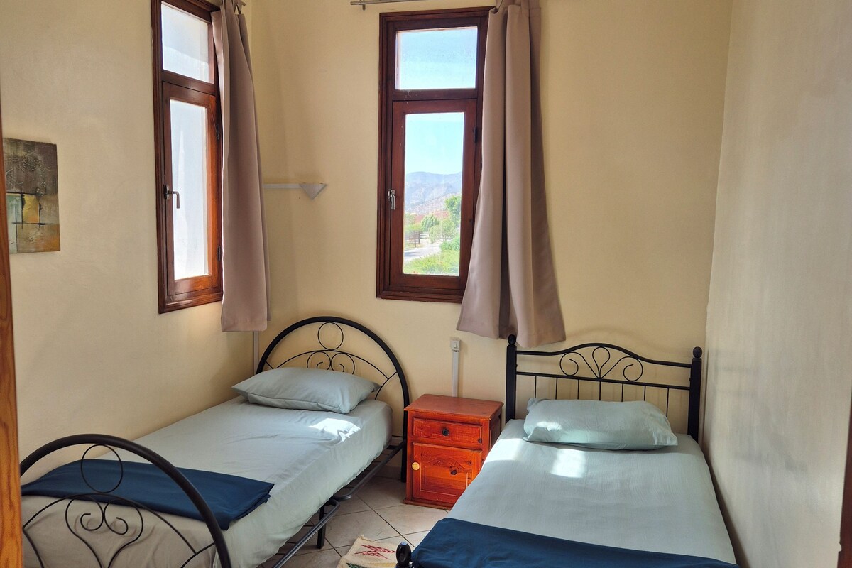 Ein Schlafzimmer mit zwei Einzelbetten, einem fensterbelaubten Fenster, einem kleinen Nachttisch aus Holz und einem Fliesenboden.