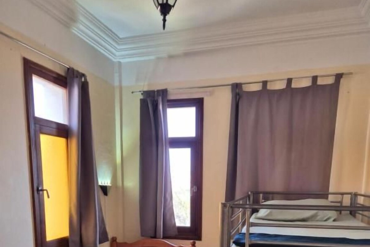 Ein Zimmer mit zwei Einzelbetten, einem Etagenbett, einem kleinen Tisch und einem Fenster mit Vorhängen.
