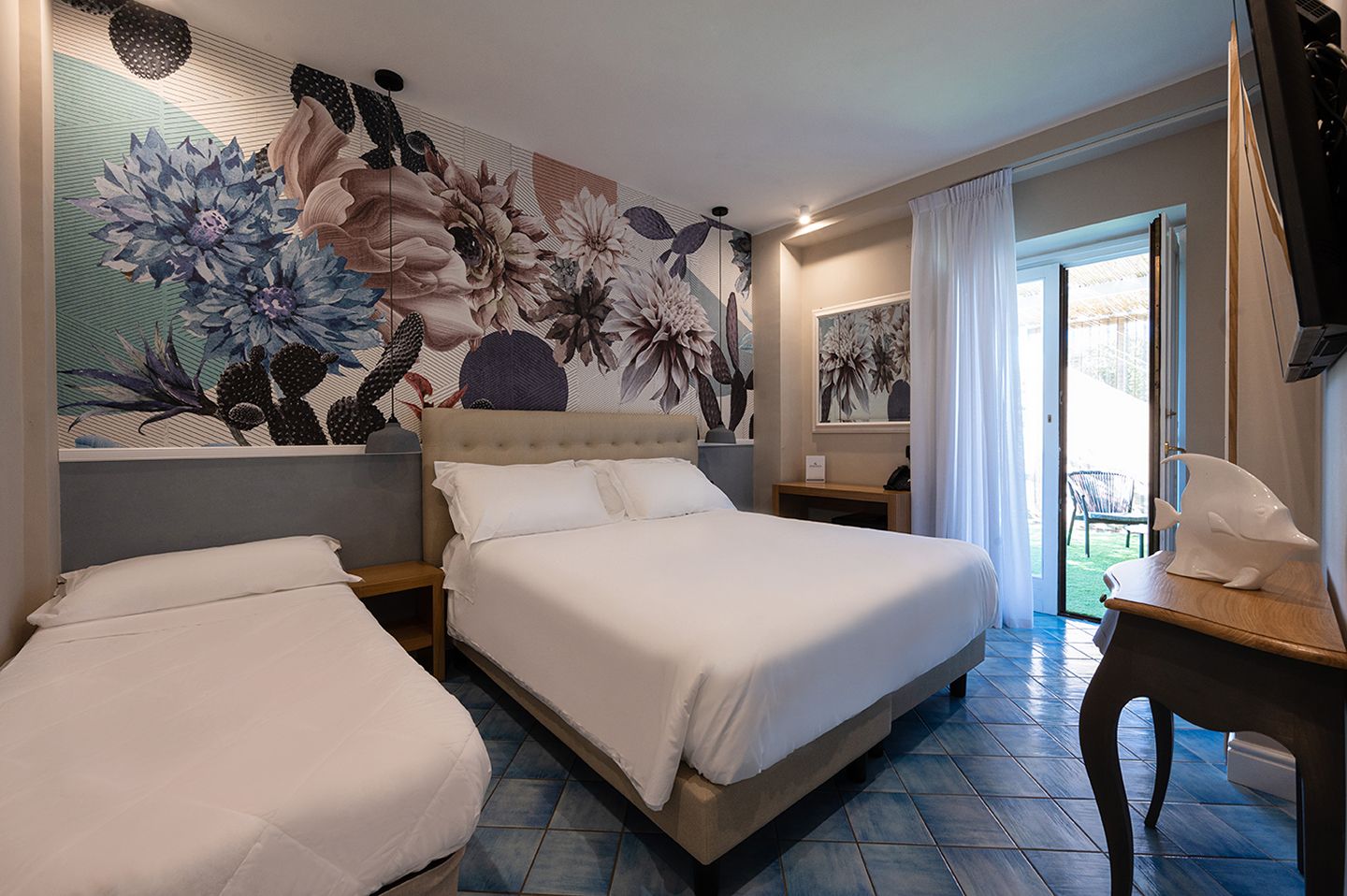 A hotel room with a large bed, a smaller bed, a desk, and a chair. There is a large floral mural on the wall and a window with curtains.