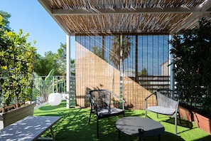 Un patio al aire libre con césped artificial, dos sillas de mimbre, una mesa redonda y un techo de bambú.