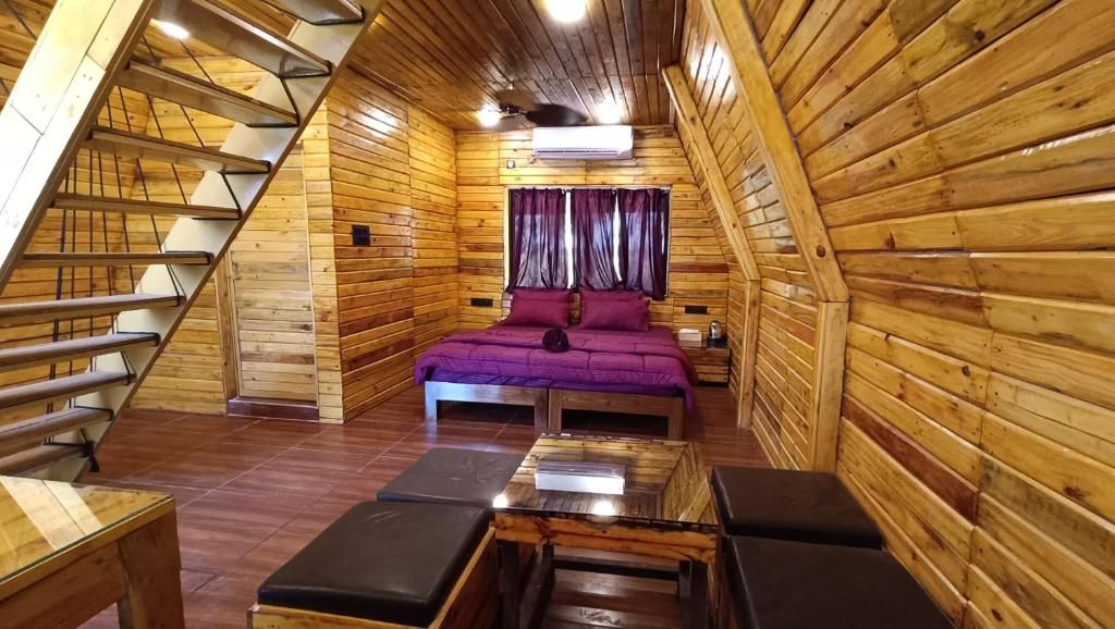 A wooden cabin with a sloped ceiling, a bed with purple bedding, a small table, and a bench.