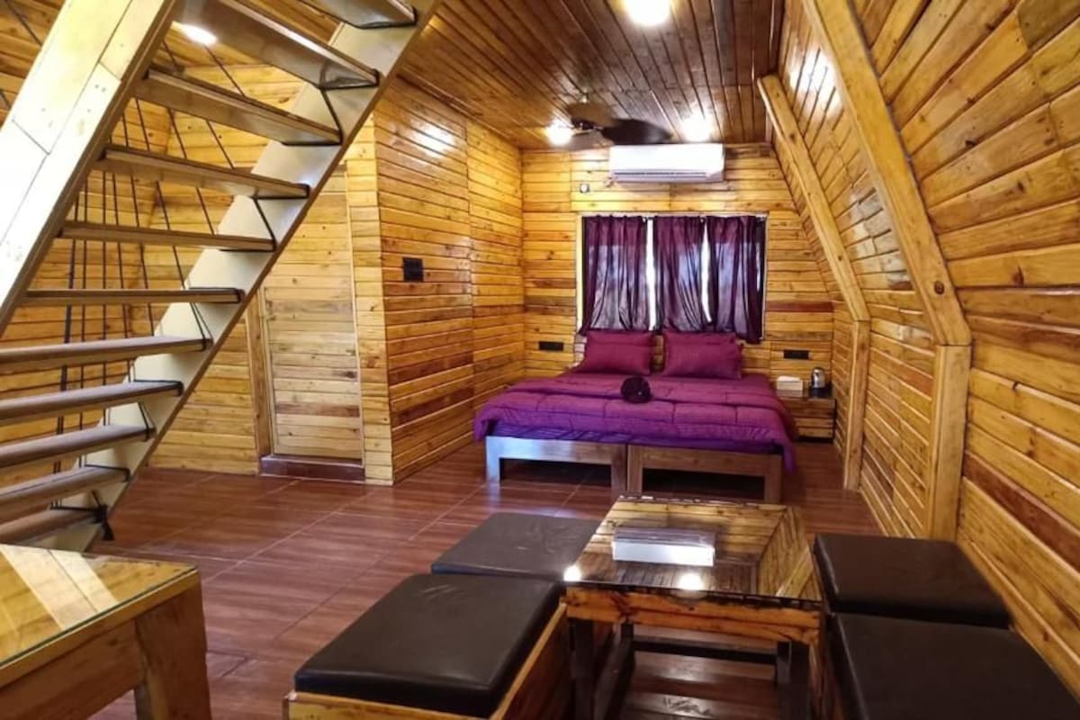 A wooden cabin with a sloped ceiling, a bed with purple bedding, a small table, and a bench.