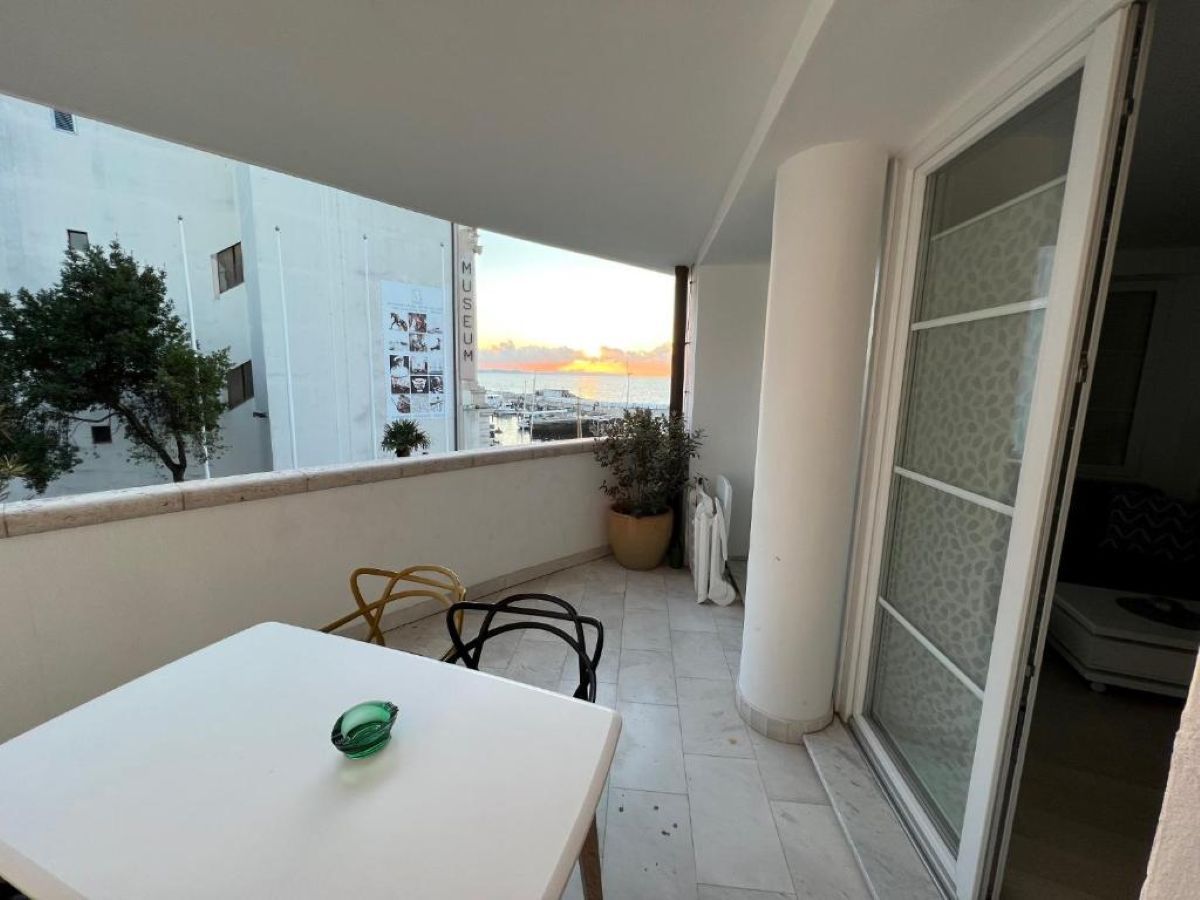 Een balkon met een witte tafel, twee stoelen, een potplant en uitzicht op zee bij zonsondergang.