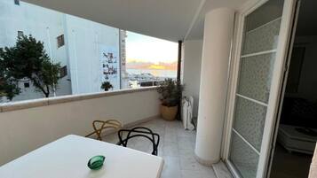 Een balkon met een witte tafel, twee stoelen, een potplant en uitzicht op zee bij zonsondergang.