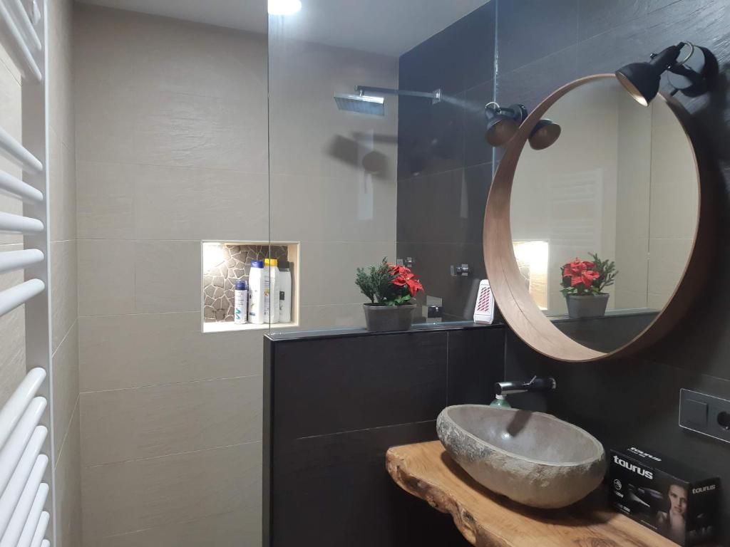 Un baño con un espejo redondo, un estante de madera, un lavabo de piedra y una pequeña ventana con plantas en macetas.