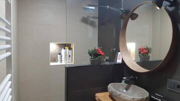 Un baño con un espejo redondo, un estante de madera, un lavabo de piedra y una pequeña ventana con plantas en macetas.