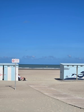 Ein Strand mit einer Reihe von nummerierten Strandkörben, einem Schild, das auf das Schwimmverbot hinweist, und einer Person, die in einem Stuhl sitzt.