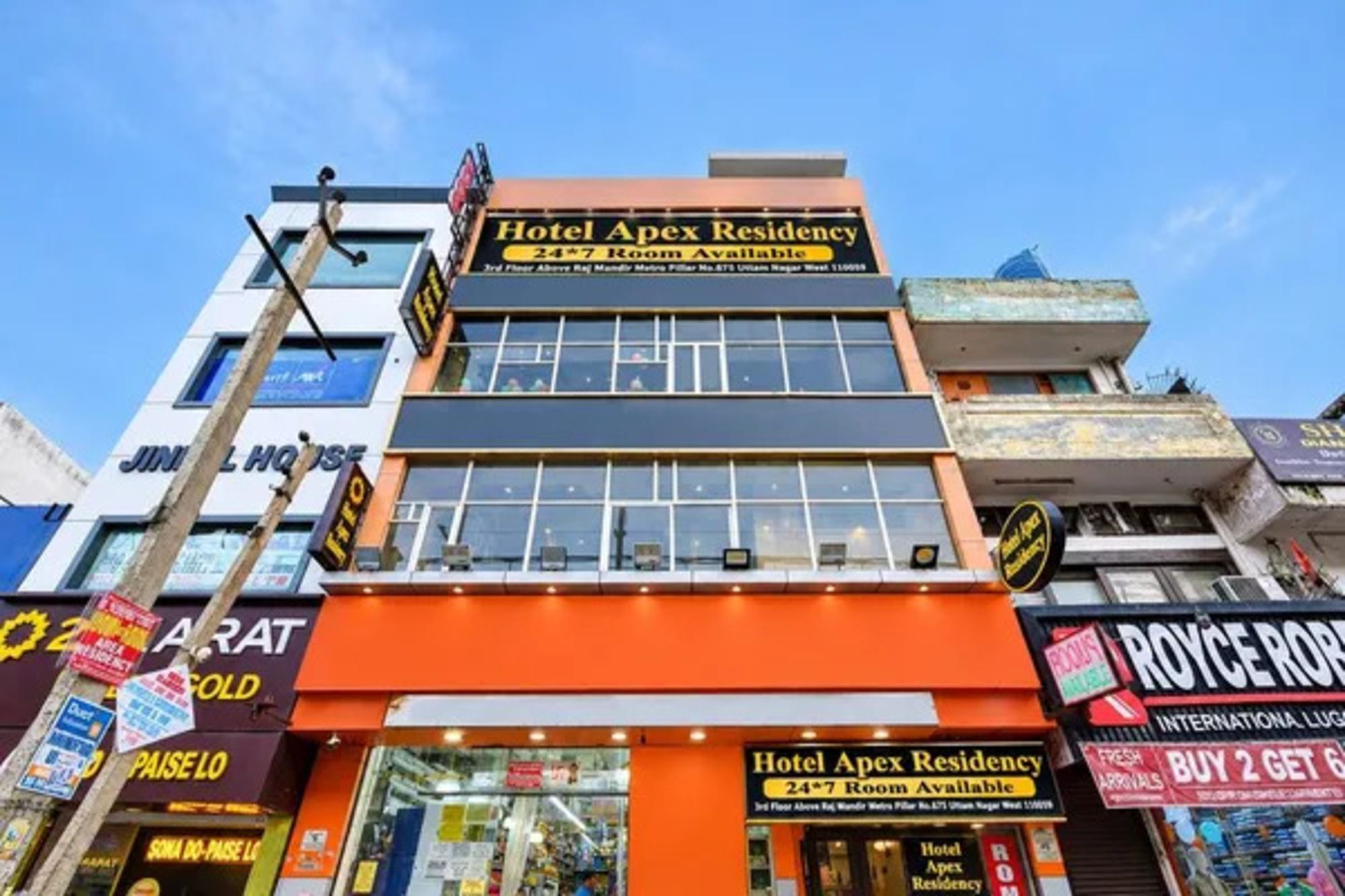 Un immeuble à plusieurs étages, sur lequel on peut voir une enseigne bien visible indiquant « Hotel Apex Residency » et précisant que les chambres sont disponibles 24 heures sur 24, 7 jours sur 7.