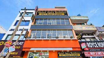 Un immeuble à plusieurs étages, sur lequel on peut voir une enseigne bien visible indiquant « Hotel Apex Residency » et précisant que les chambres sont disponibles 24 heures sur 24, 7 jours sur 7.