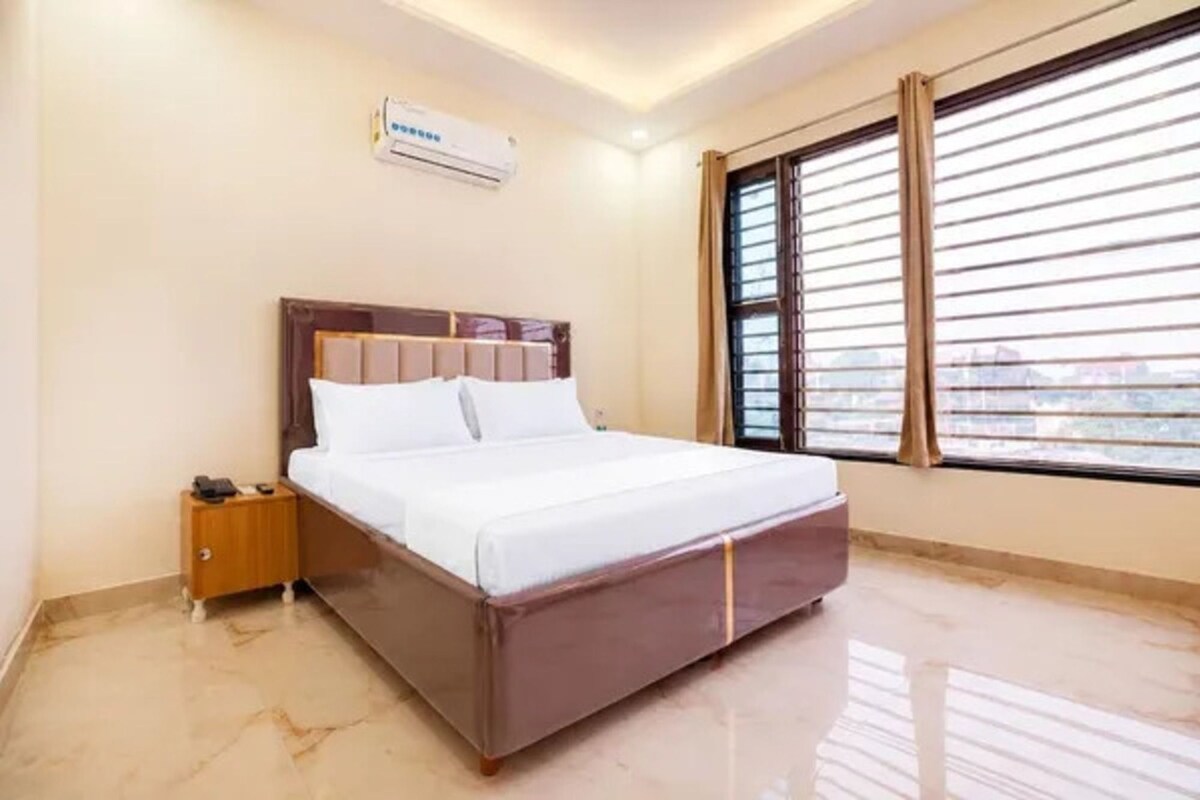 Una habitación de hotel con cama, mesita de noche, equipo de aire acondicionado y ventana con persianas.