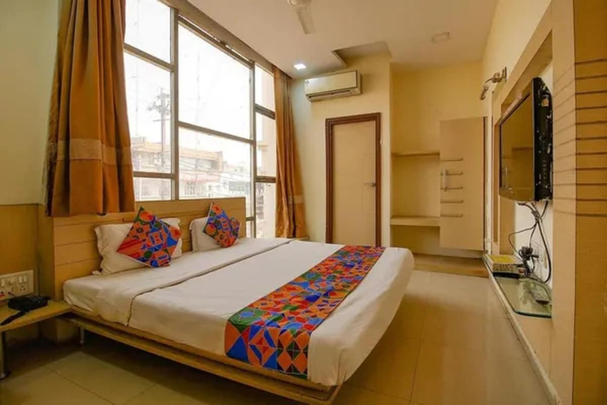 A hotel room with a bed, a bedside table, an air conditioning unit, a mirror, and a window with curtains.