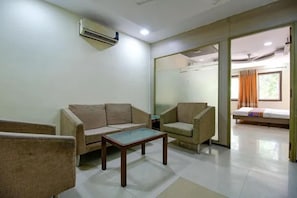 A room with a sofa, armchair, and a small table. There is a bed with a window and curtains in the background. - FabHotel Maanpreet (Bhopal)