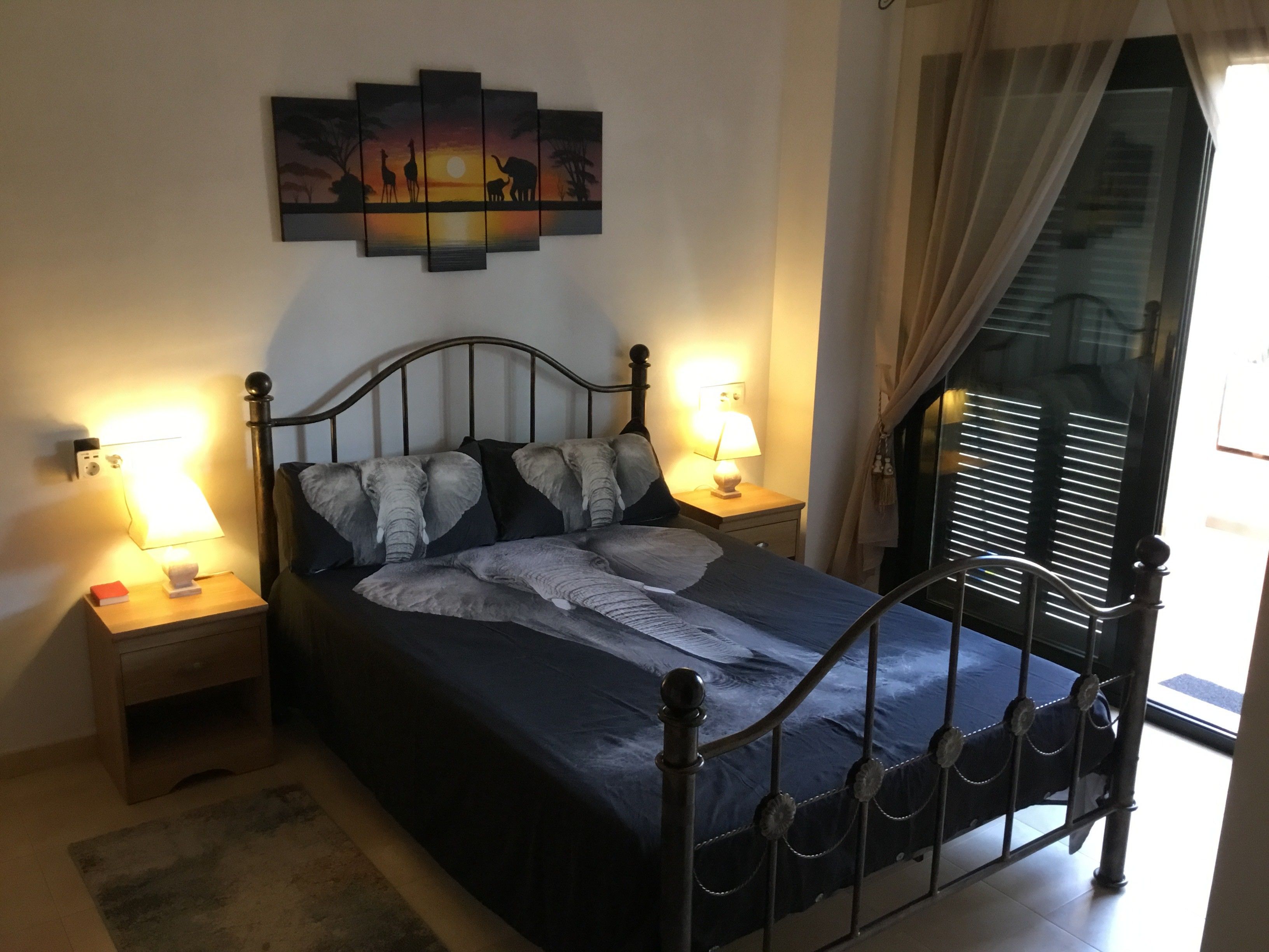 Una cama con estructura metálica, dos mesitas de noche con lámparas, una obra de arte en la pared y una ventana con cortinas.