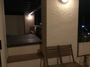 Un'area esterna scarsamente illuminata con una panca di legno, un muro dalla superficie strutturata e un piccolo fiorile.