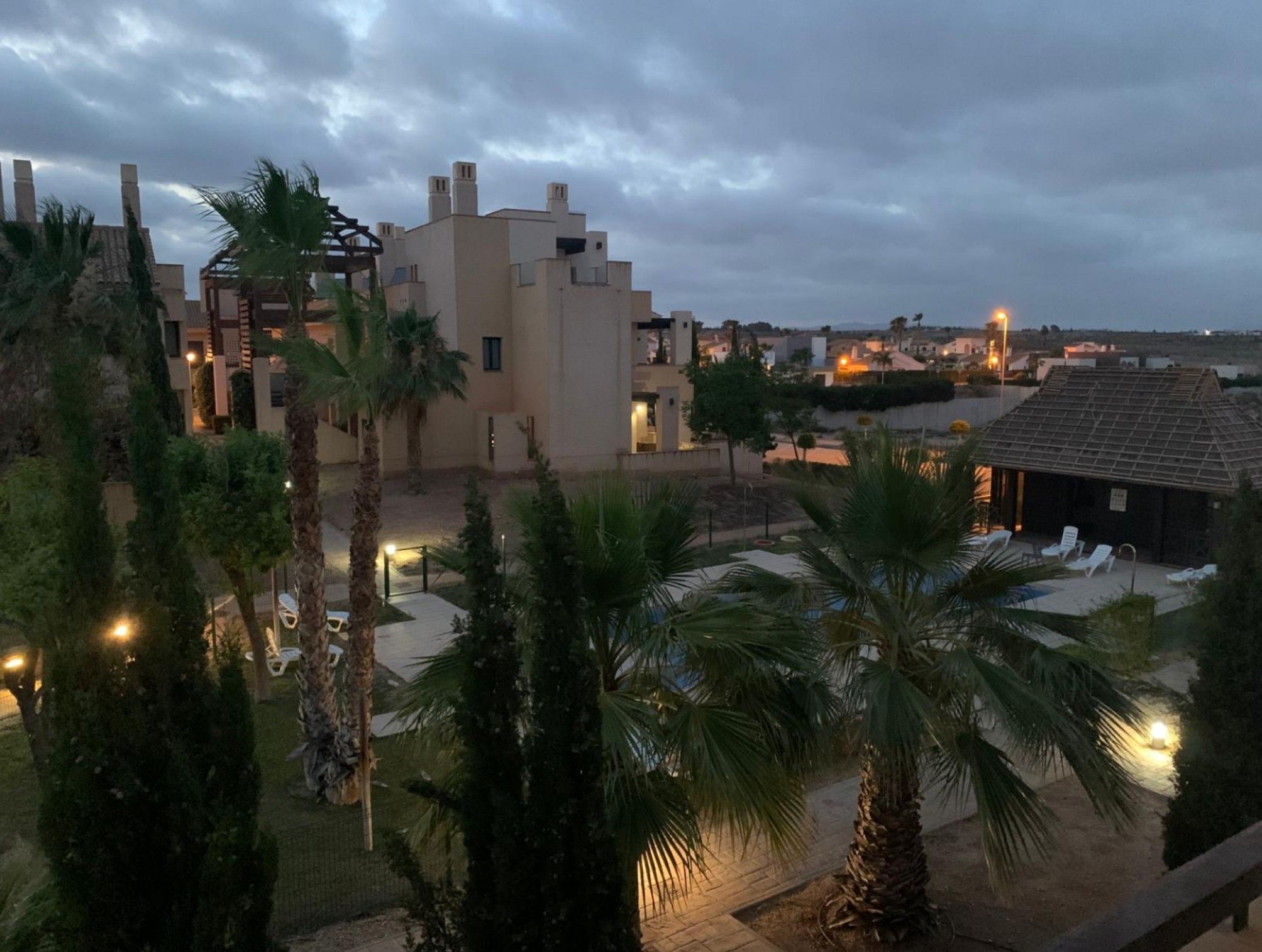 A residential area with modern architecture, including a building with a flat roof and a balcony. There are palm trees and a paved area with lounge chairs.
