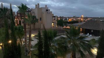 A residential area with modern architecture, including a building with a flat roof and a balcony. There are palm trees and a paved area with lounge chairs.