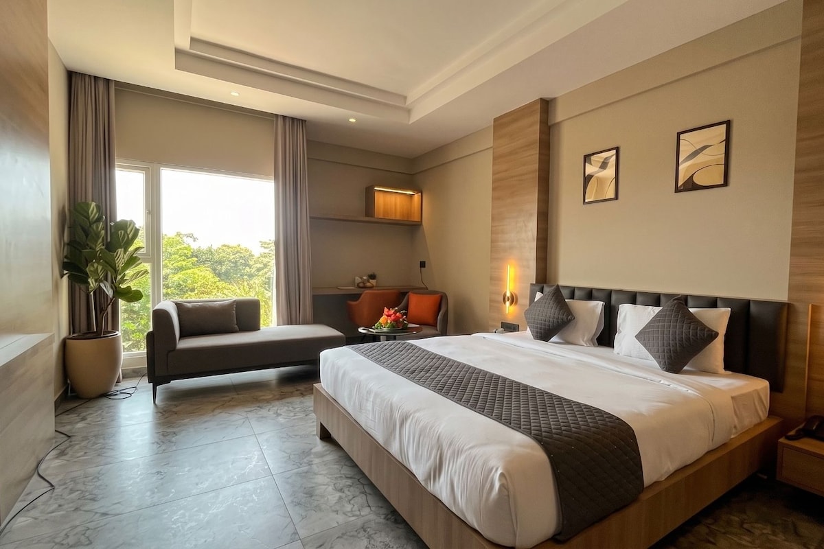 A modern hotel room with a large bed, a sofa, a desk, and a view of greenery outside.