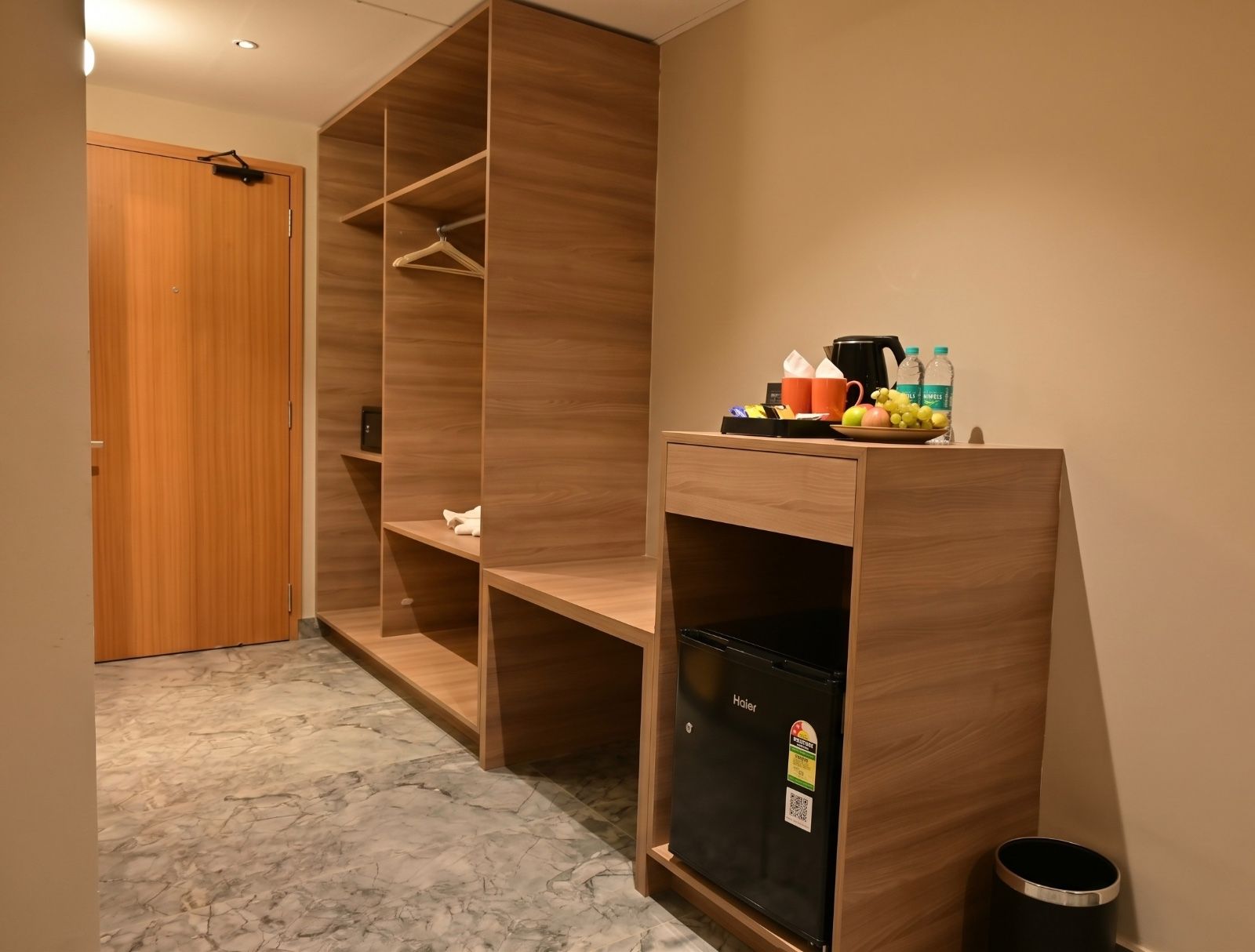 Une chambre d’hôtel avec une armoire en bois, un bureau sur lequel se trouve un petit réfrigérateur noir, et une petite poubelle.