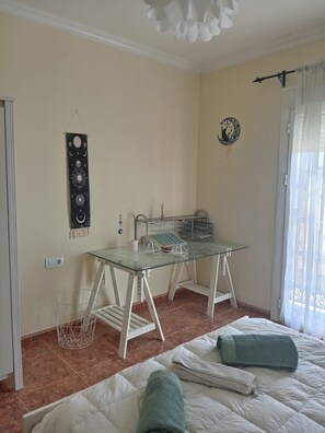 Una habitación con una mesa de vidrio, una cama con una toalla verde, un perchero de pared y una lámpara en el techo.