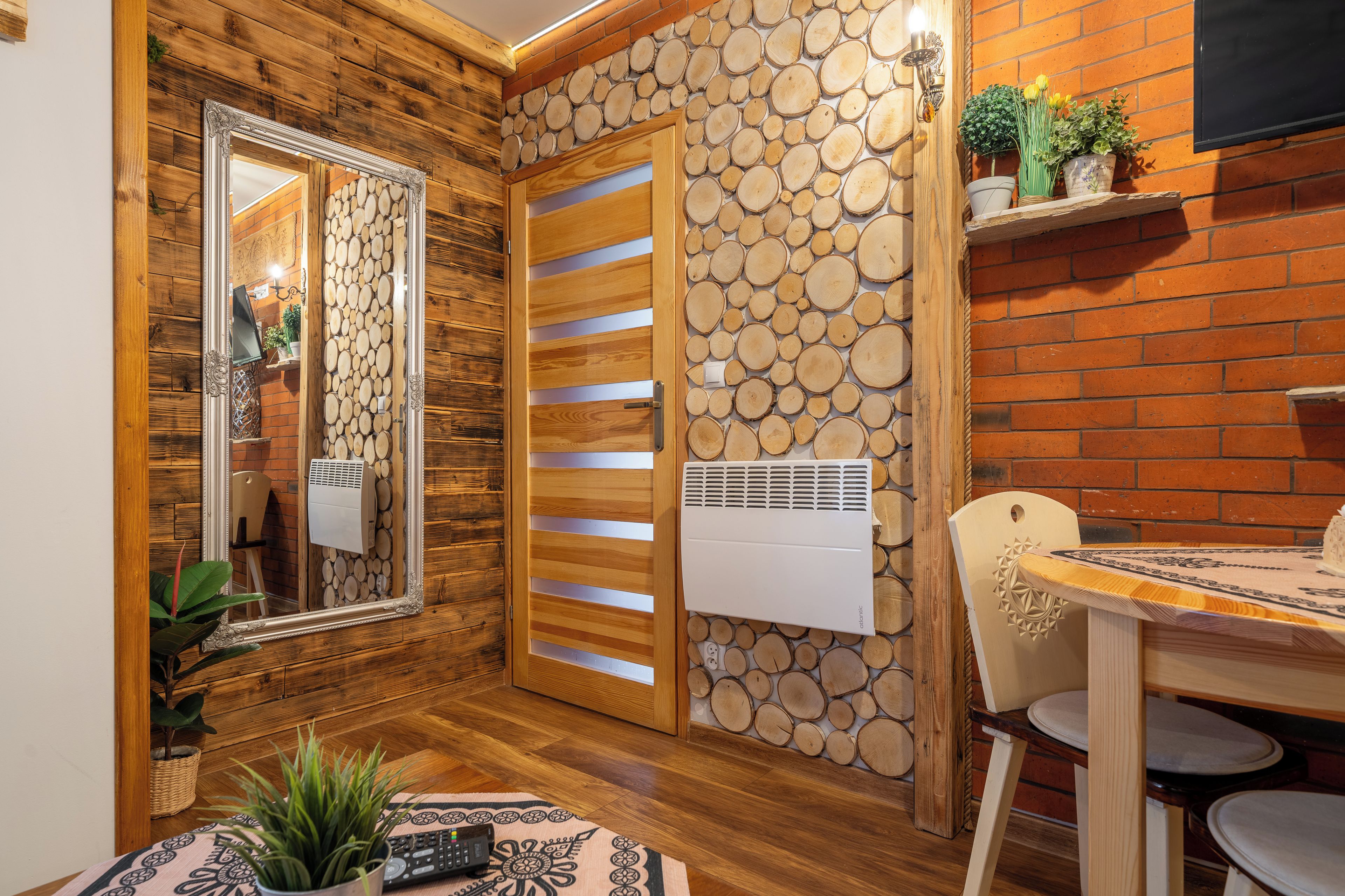 A room with a wooden floor, a mirror, a door with a wooden panel, a wall with a log pattern, a table with chairs, and a potted plant.
