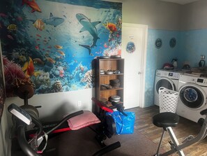 Habitación con un gran mural de una escena submarina, una caminadora, una bicicleta estática, una lavadora y un estante con varios objetos.