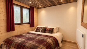 A bedroom with a wooden ceiling, a bed with a patterned blanket, red curtains, and a window with blinds. - New Listing (Châtel)