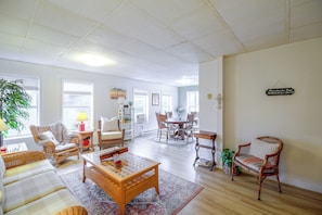 A spacious living room with a wooden coffee table, wicker chairs, a dining table with chairs, and a large window providing natural light.