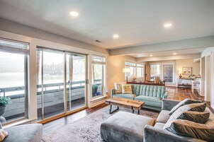 A spacious living room with a large window overlooking a body of water, a cozy seating area with a sofa and ottoman, and a kitchen area in the background.
