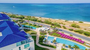 Aerial view of beachfront property featuring pool facilities, garden areas, and oceanfront road adjacent to sandy shoreline with turquoise waters. - 1BR Retreat for Two | Stunning 6th-Floor Views by BlueAruba (Noord)