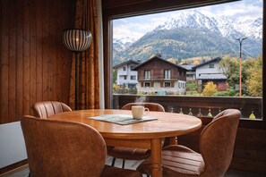 A wooden interior with a round table and chairs, a window with a view of houses and mountains. - New Listing (Les Houches)