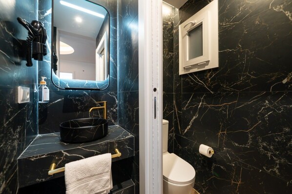 Un baño con lavamanos de mármol negro, detalles dorados, un inodoro blanco y un espejo pequeño.