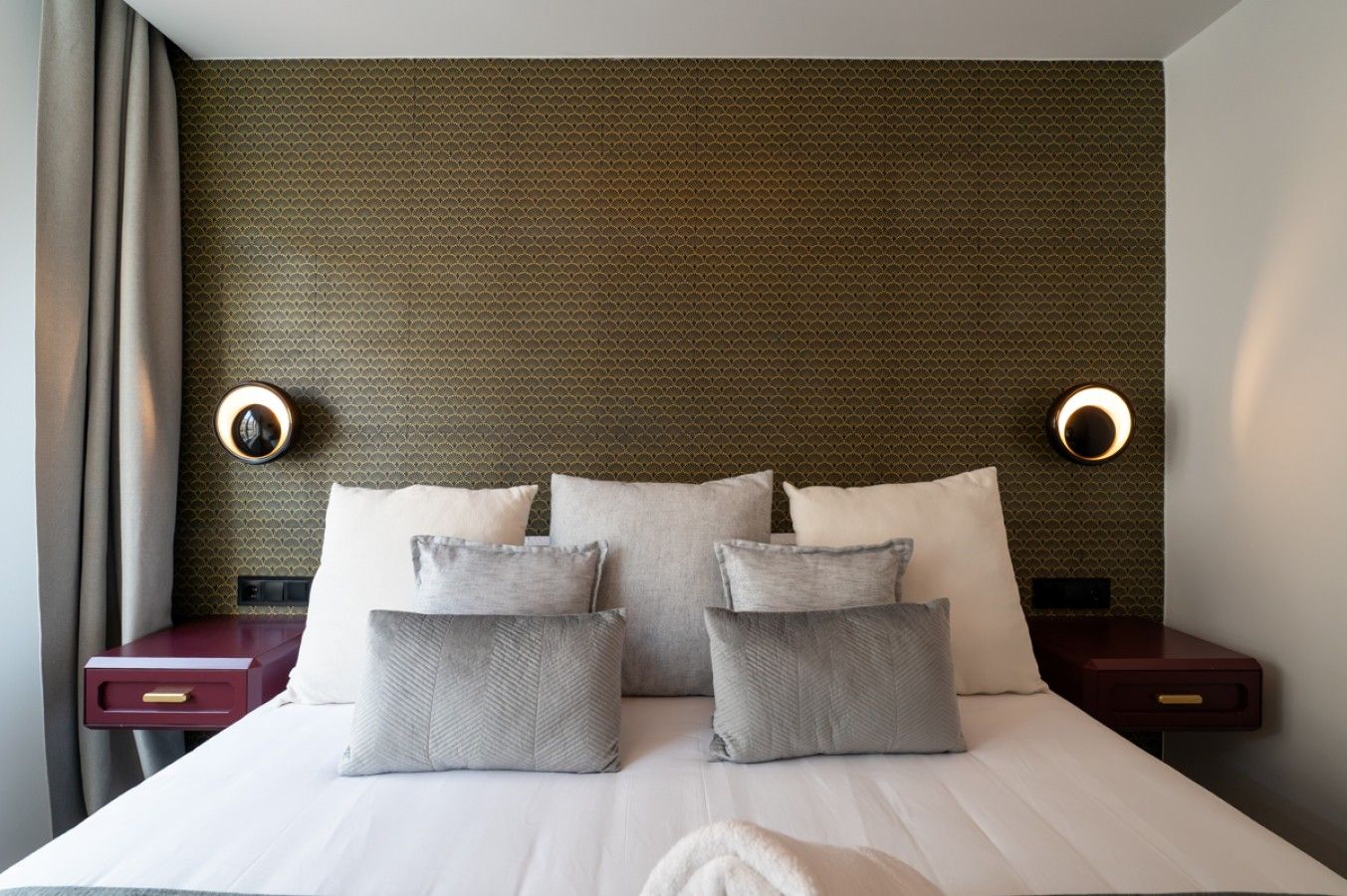 A hotel room with a bed, pillows, a nightstand, and wall sconces.