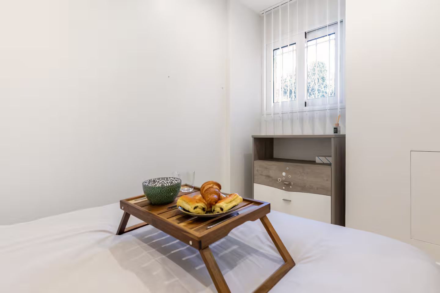 Een slaapkamer met een bed, een houten nachtkastje met een dienblad waarop een kom, een bord met croissants en een raam met gordijnen staan.