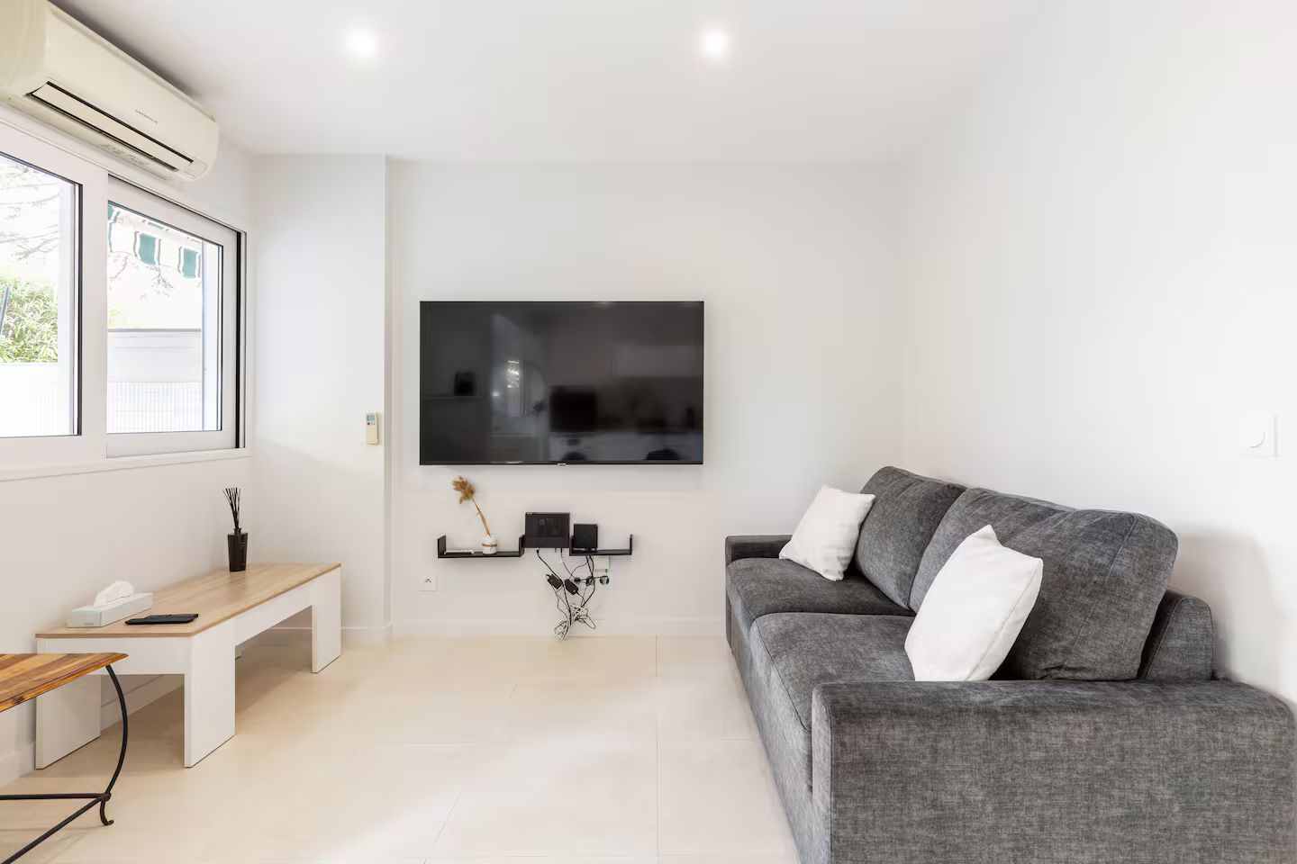 Een moderne woonkamer met een grijze bank, een flatscreen televisie aan de muur, een houten bankje en een kleine tafel met een vaas en een afstandsbediening.