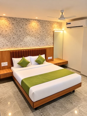 Une chambre d’hôtel moderne avec une tête de lit en bois, un grand lit avec une literie blanche et des touches de vert, une table de chevet, un miroir et un appareil de climatisation.