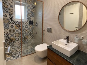 A bathroom with a patterned tile wall, a white toilet, a sink with a mirror, and a shower with a glass door. - SERA LUXURY apartment in quaint Noida (Noida)