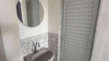 A bathroom with a pedestal sink, a round mirror, and a door with horizontal blinds.