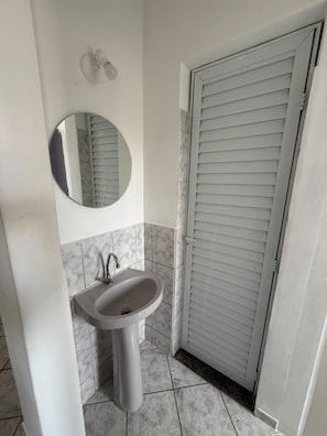 A bathroom with a pedestal sink, a round mirror, and a door with horizontal blinds. - Apartamento aconchegante (Vitória)