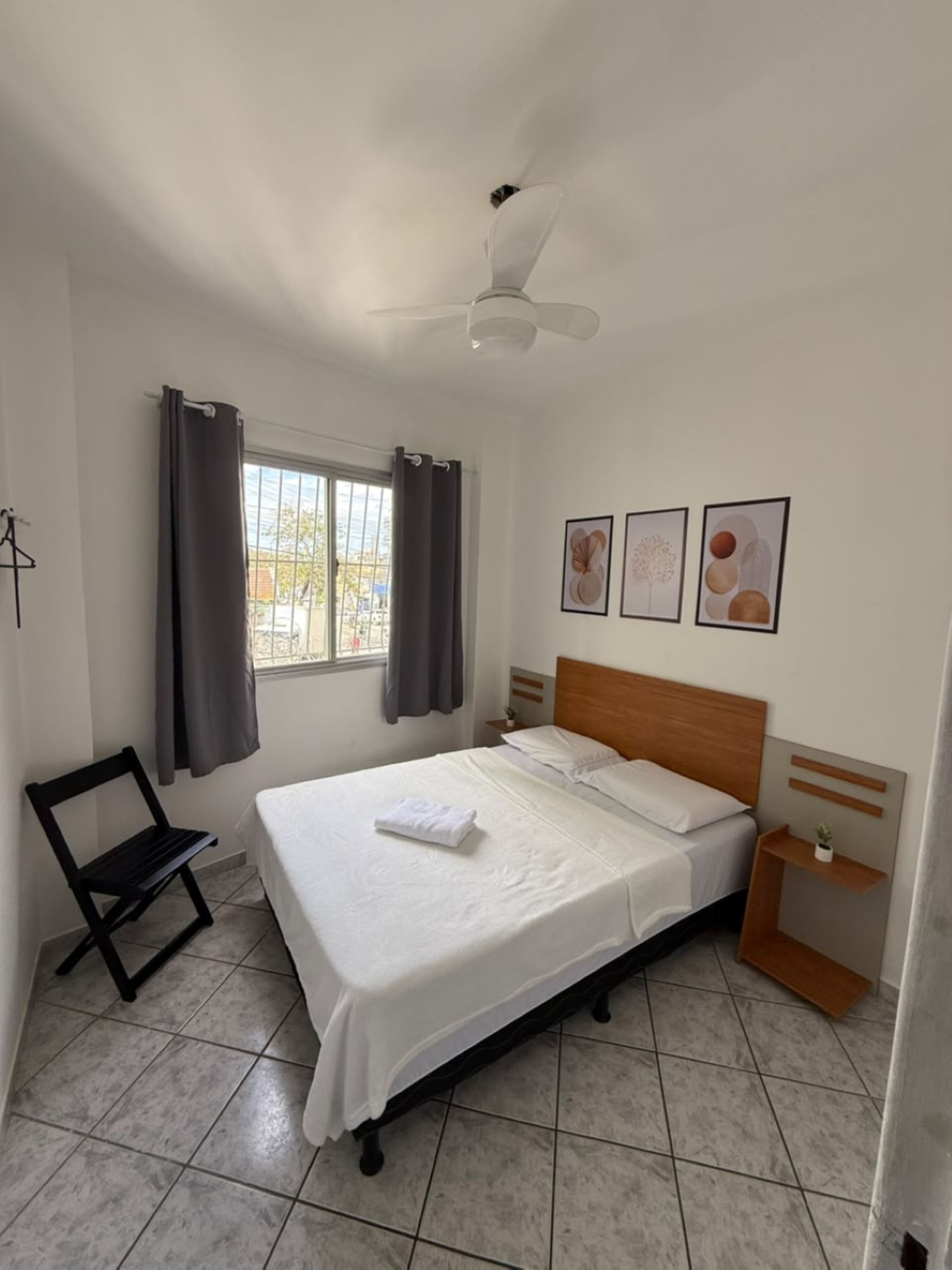 A bedroom with a bed, a nightstand, a chair, a window with curtains, and wall art.