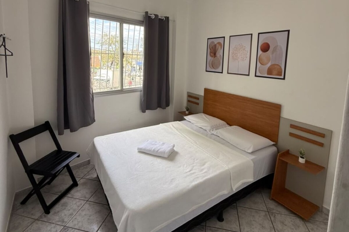 A bedroom with a bed, a nightstand, a chair, a window with curtains, and wall art.