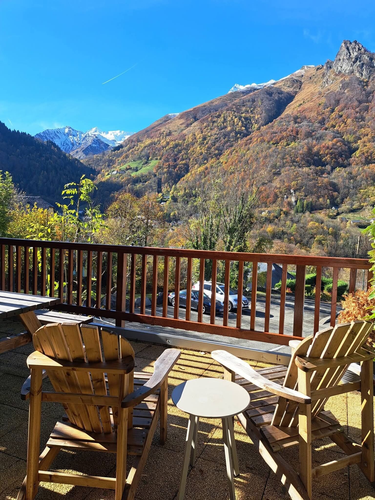 Une terrasse en bois, dotée de chaises Adirondack et d’une table, offre une vue imprenable sur un paysage montagneux sous un ciel dégagé.