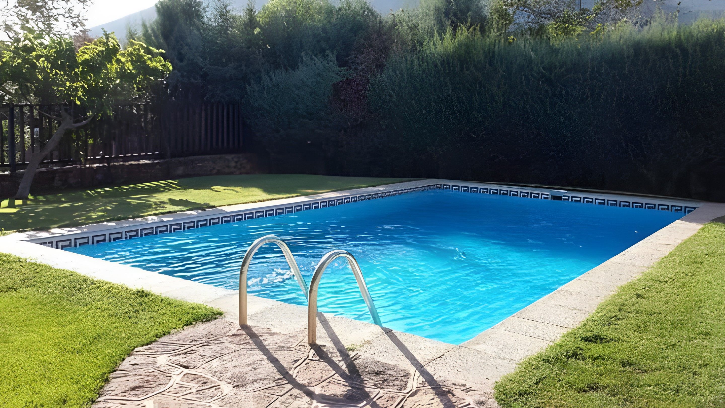 Una piscina rectangular con superficie de agua azul cristalina, rodeada por una solana de piedra y una zona de césped.