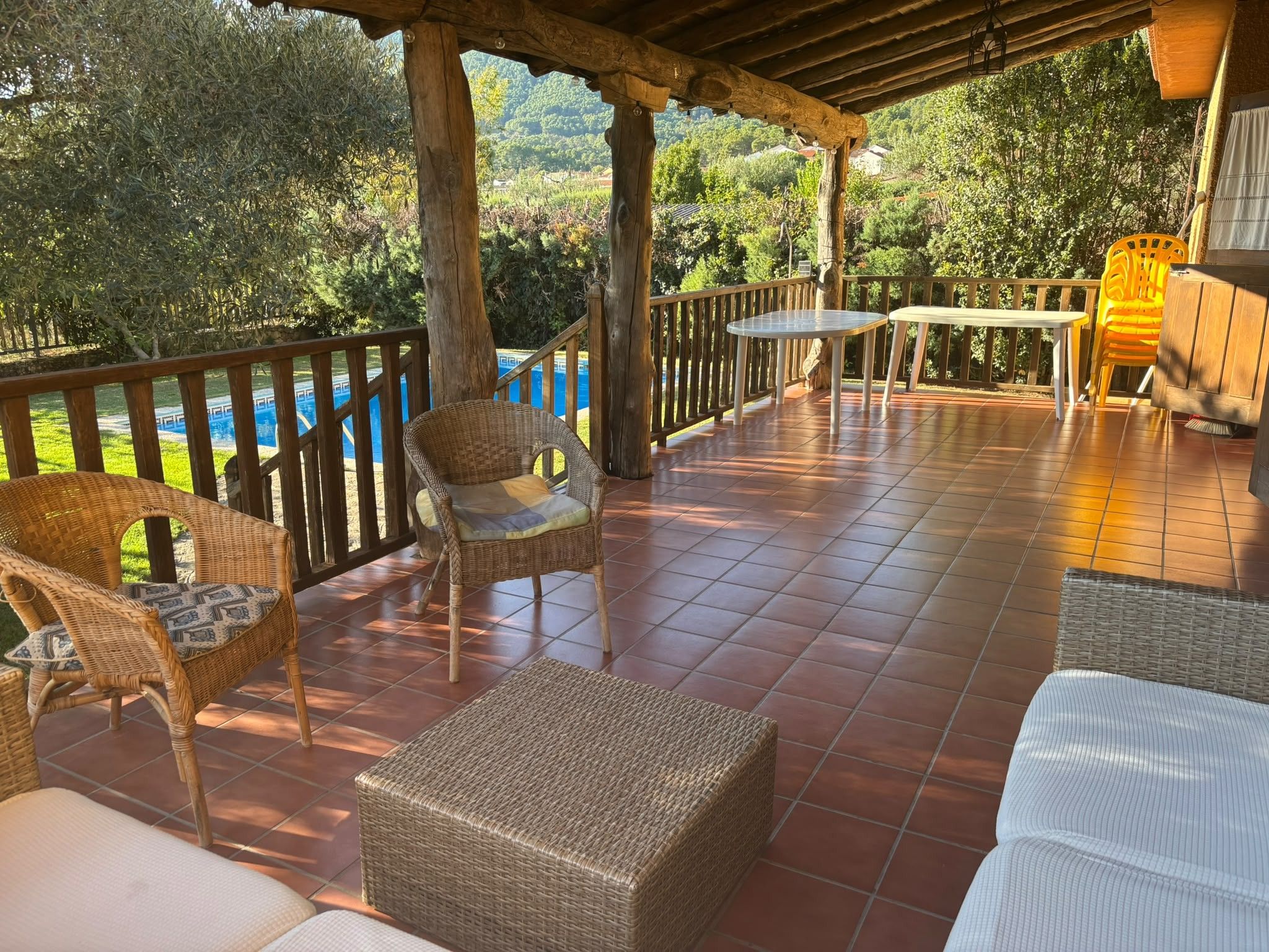 Una veranda coperta con mobili in vimini, un tavolo e vista su una piscina e vegetazione.