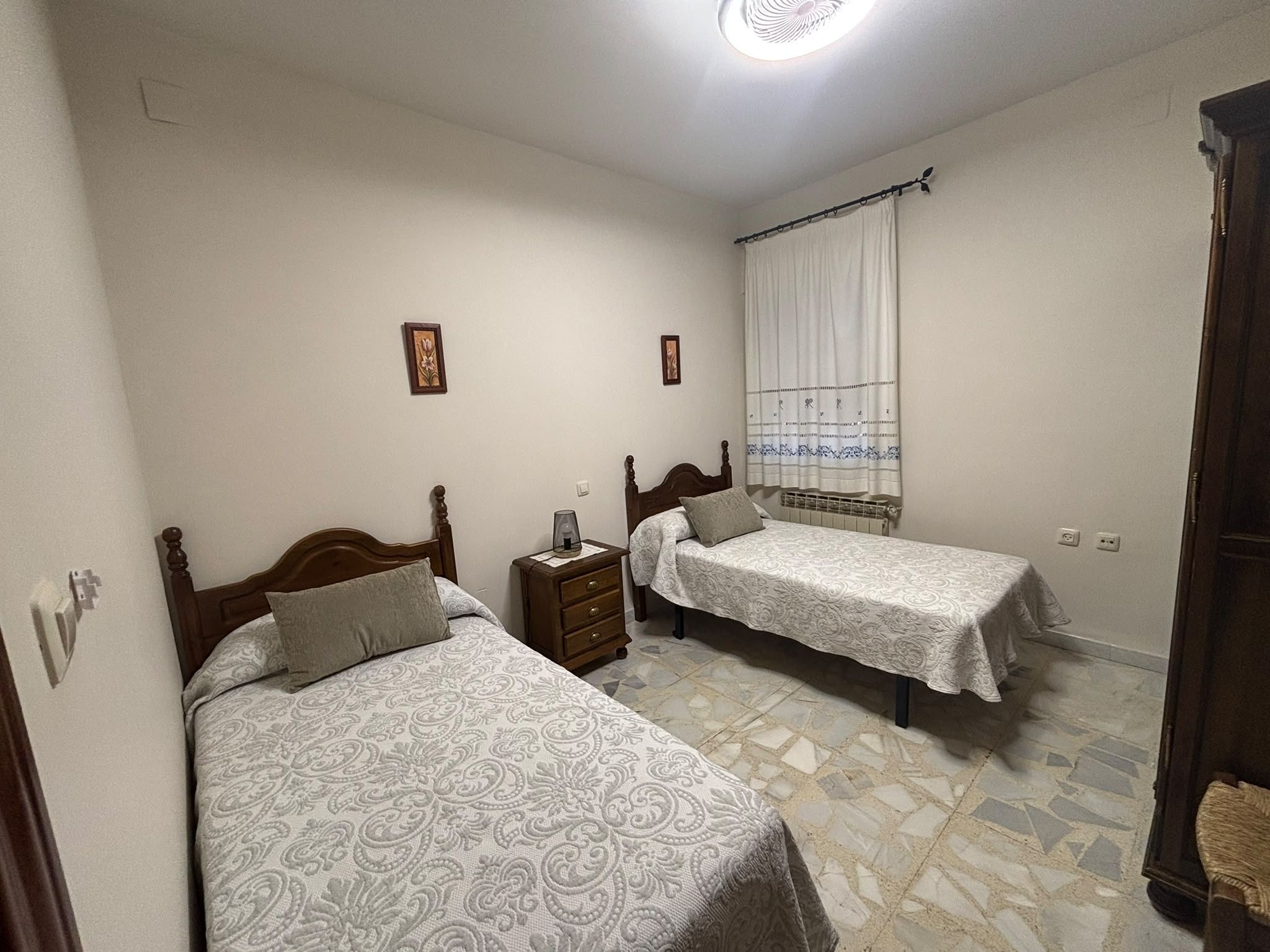 Un dormitorio con dos camas individuales, una mesita de noche, una ventana con cortinas y un radiador.