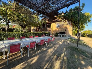 Un área de comedor al aire libre con una mesa larga y sillas rojas, rodeada de árboles y un edificio con balcón.