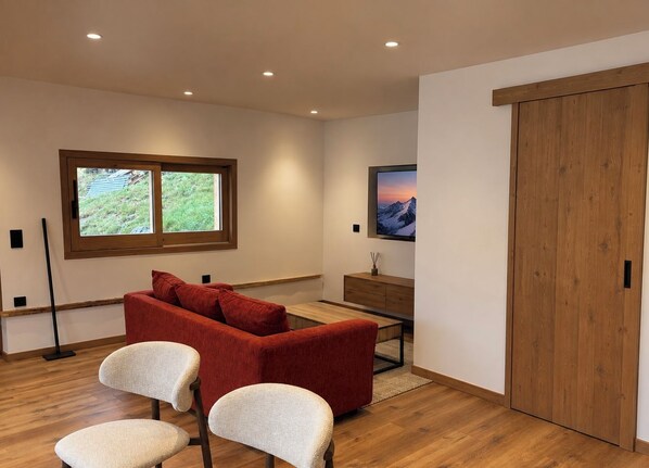 Un salon moderne avec un canapé rouge, deux fauteuils blancs, une table basse en bois, un téléviseur fixé au mur et une porte coulissante en bois.