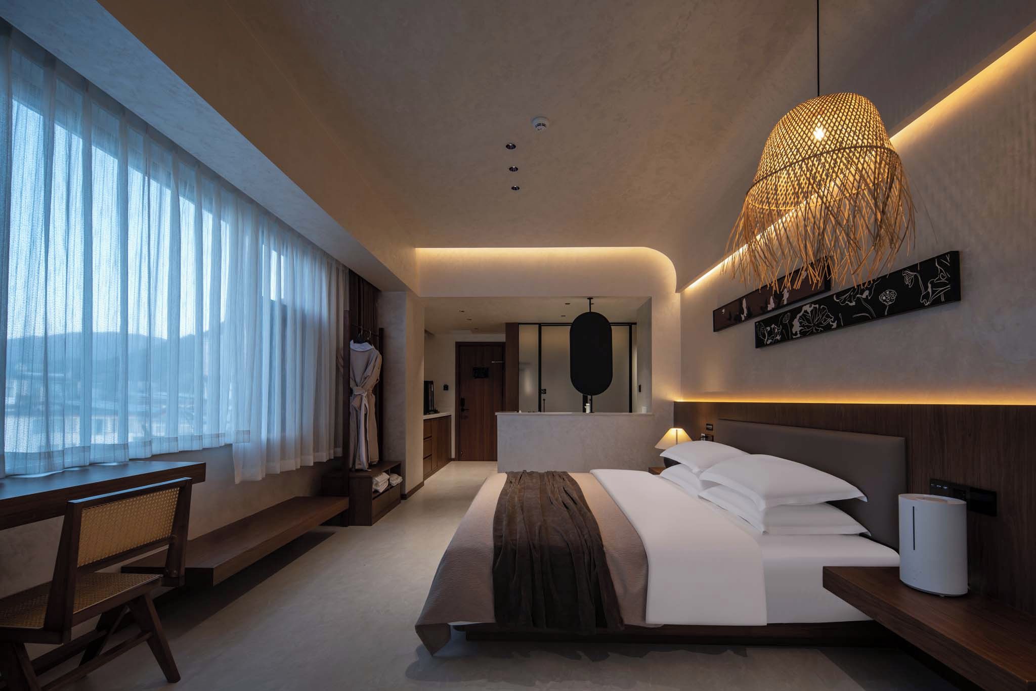 Une chambre d’hôtel moderne avec un grand lit, un bureau et une vue sur la ville à travers de fines rideaux.