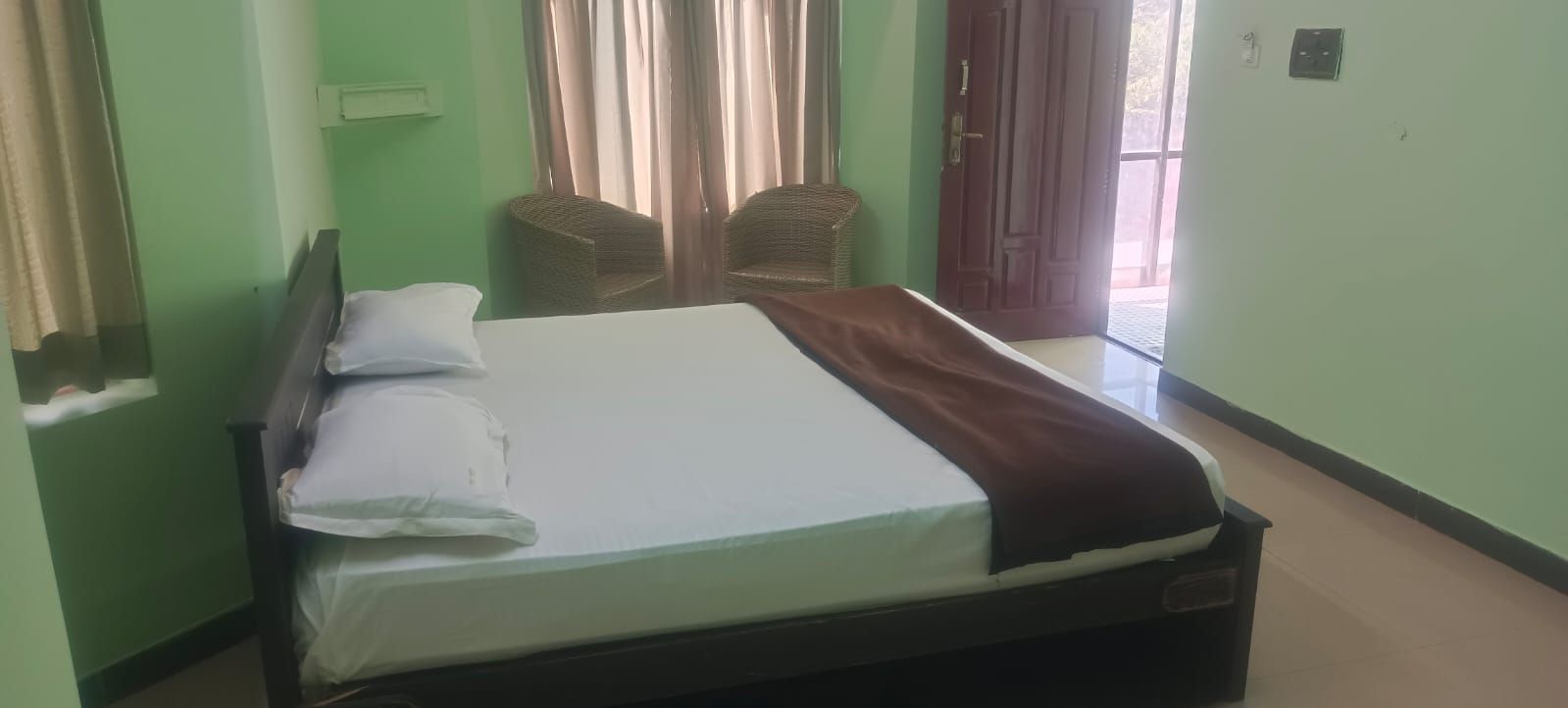Una cama individual con almohadas y una manta, dos sillas de mimbre, una puerta y un interruptor de luz en la pared.