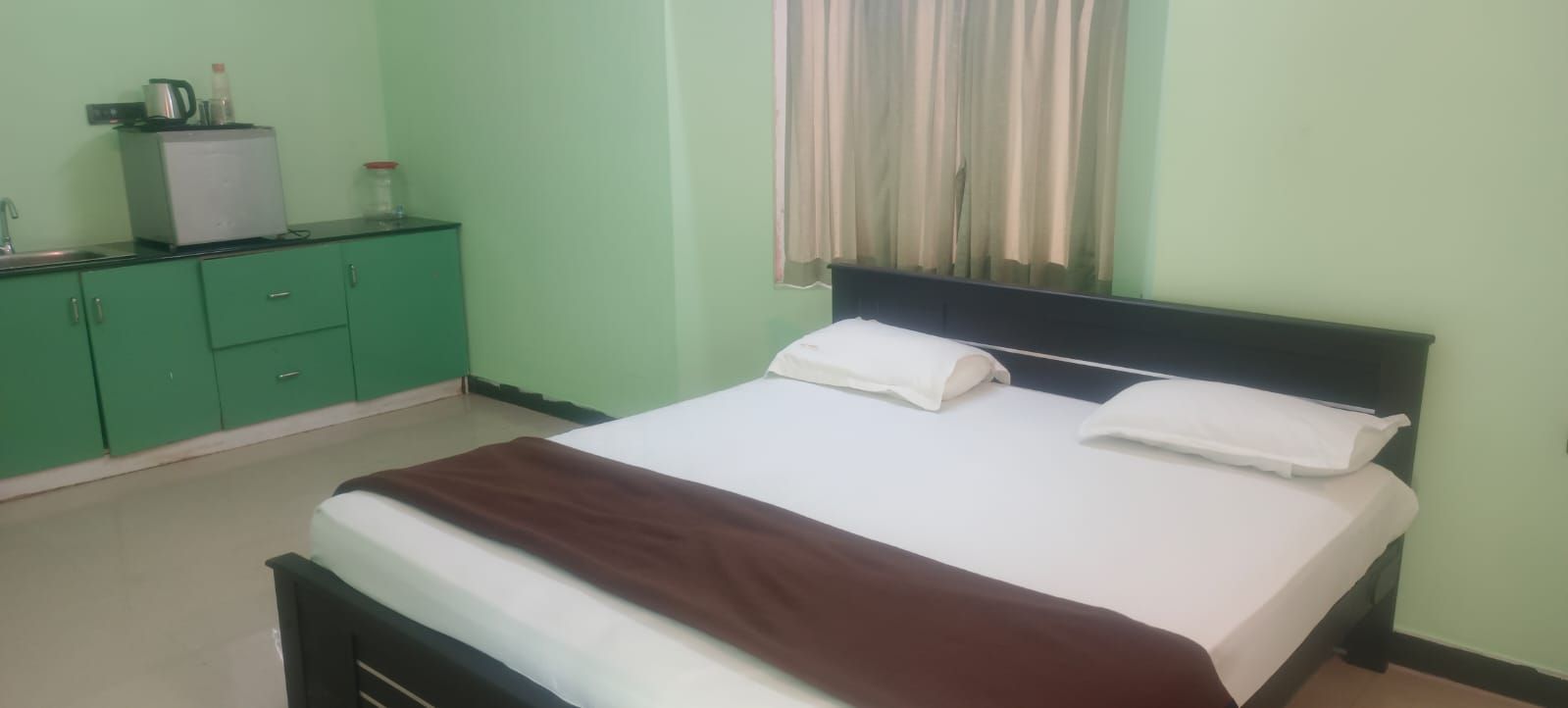 A room with a bed, a green dresser, a small refrigerator, and a window with curtains.