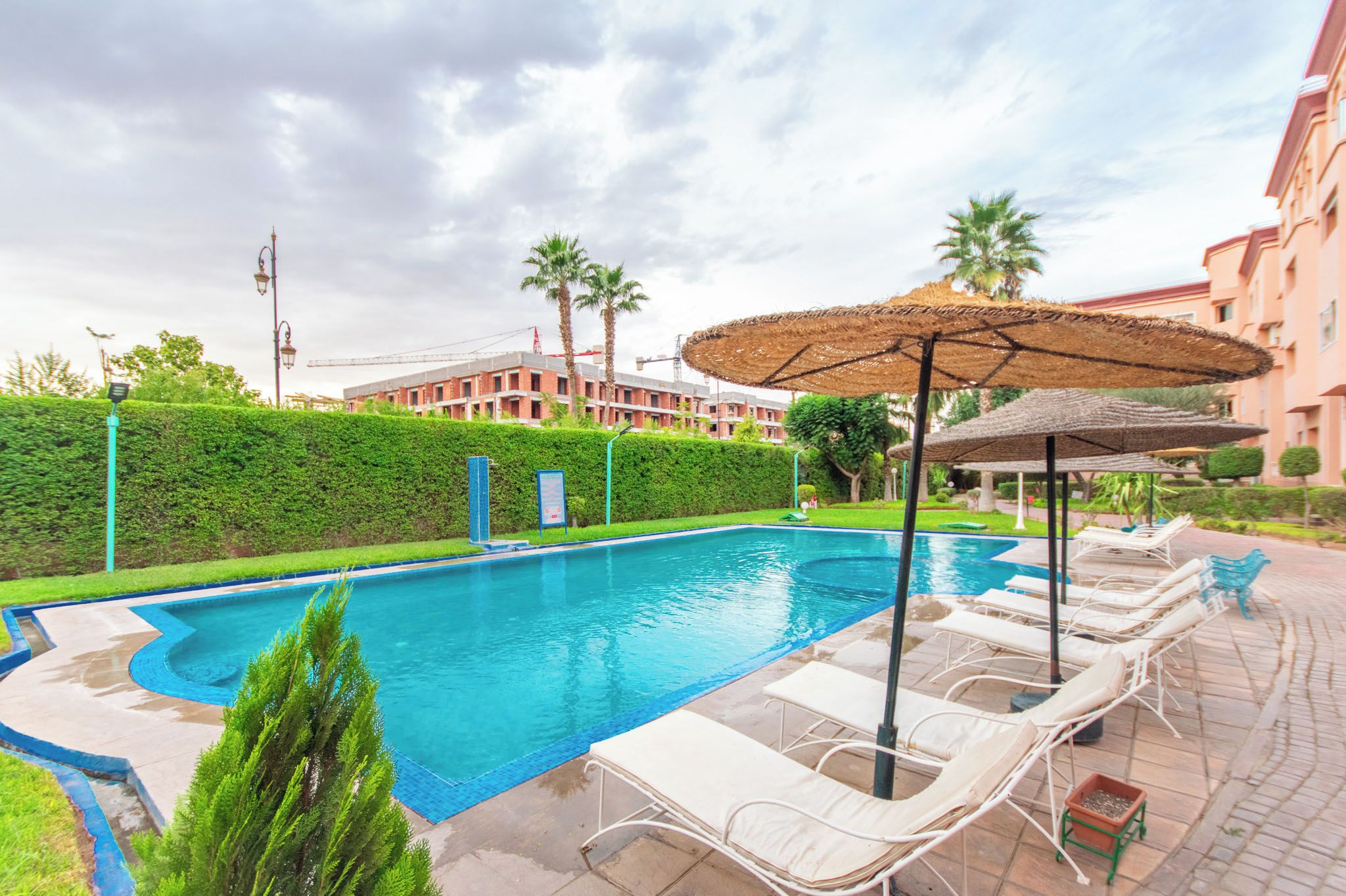 A swimming pool with lounge chairs and umbrellas, surrounded by a hedge and a building in the background.